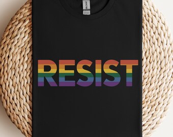 May include: Black t-shirt with the word "RESIST" in rainbow colors. The shirt is folded and placed on a round, woven, light brown surface. The text is in a bold, sans-serif font.