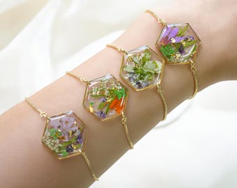 Könnte beinhalten: Vier goldfarbene Armbänder mit sechseckigen Anhängern, gefüllt mit getrockneten Blumen in verschiedenen Farben. Die Blumen, darunter lila, orange, weiß und grün, sind in klarem Harz eingeschlossen. Die Armbänder sind durch feine Goldketten verbunden.