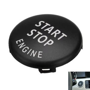 Op de afbeelding: Een ronde zwarte start-stopknop voor de motor met de witte tekst START STOP ENGINE op het oppervlak. De knop heeft een gladde afwerking en is voorzien van een klein bevestigingslipje aan de onderrand voor installatie in een voertuigdashboard.
