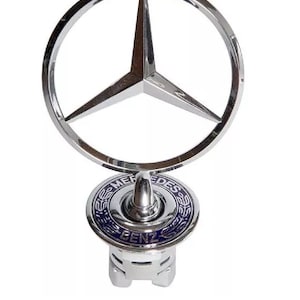 May include: A chrome Mercedes-Benz hood ornament featuring the iconic three-pointed star within a circular frame. The base has a blue and silver emblem with the words "Mercedes Benz".