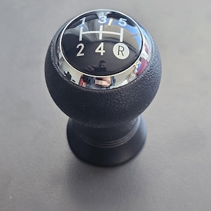 Może przedstawiać: Czarny skórzany gałka zmiany biegów z chromowanym pierścieniem i białymi cyframi 1, 2, 3, 4, 5 i R dla biegu wstecznego.