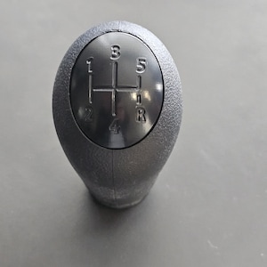May include: Black plastic gear shift knob with a 5-speed pattern, including reverse, labeled 1, 2, 3, 4, 5, and R.