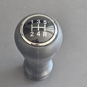 Può includere: Un pomello del cambio nero con un anello cromato e uno schema di marcia nero con numeri bianchi 1, 3, 5, 2, 4 e R.