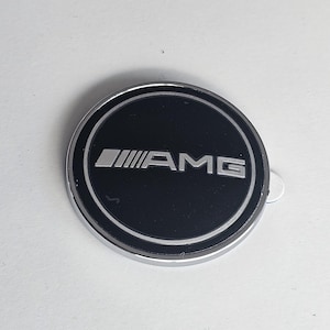 Puede incluir: Un emblema redondo de color plateado con fondo negro. El emblema tiene la palabra "AMG" en letras plateadas.