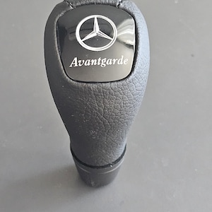 Op de afbeelding: Zwarte leren versnellingspookknop met een zilveren Mercedes-Benz-logo en het woord "Avantgarde" erop gegraveerd.