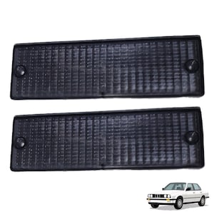 Puede incluir: Dos luces de posición laterales rectangulares negras para coche con superficie texturizada. Las luces tienen esquinas redondeadas y herrajes de montaje. Un coche blanco se muestra en la esquina inferior derecha.
