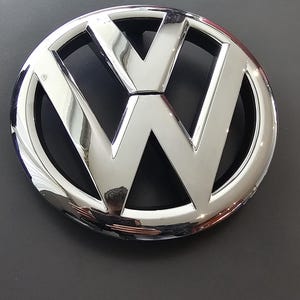 Peut inclure: Un emblème Volkswagen chromé, avec un design circulaire et les lettres "V" et "W" entrelacées. L'emblème a une surface brillante et réfléchissante, sur fond sombre.