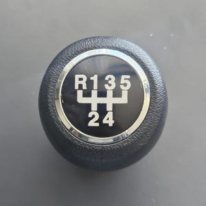 Pode incluir: Botão de mudança de marcha preto com um anel prateado e um padrão de marcha preto e branco com os números R, 1, 3, 5, 2 e 4.