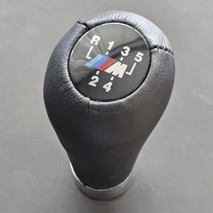 Op de afbeelding: Zwarte leren versnellingspookknop met een zilveren ring aan de basis. De knop heeft een zwarte en witte ronde inzet met de letters "R", "1", "3", "5", "L", "2", "4" en "M" erop gedrukt. De "M" is een BMW-logo.