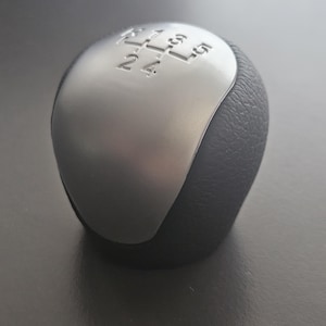 May include: A silver and black gear shift knob with a 5-speed pattern. The knob has a smooth, silver top and a textured, black bottom.