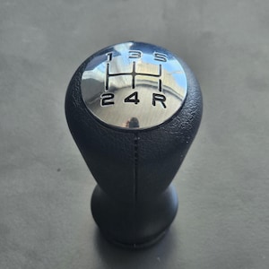 Könnte beinhalten: Ein schwarzer Leder-Schalthebelknauf mit einer silbernen Oberseite. Die Oberseite hat die Zahlen 1, 2, 3, 4, 5 und R aufgedruckt.