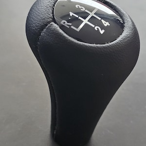 Może przedstawiać: Czarny skórzany gałka zmiany biegów z czarną plastikową górą z srebrnym wzorem zębatki i numerami "R 1 3" i "2 4".