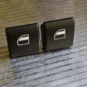 Könnte beinhalten: Zwei schwarze Kunststoff-Fensterschalter für Autos mit einem weißen Fenstersymbol auf jedem.