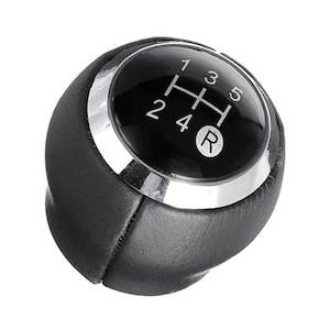 May include: A black leather gear shift knob featuring a circular top insert with a glossy black finish. The insert displays a white gear pattern for a five-speed manual transmission, including positions 1, 2, 3, 4, 5, and R, surrounded by a silver ring.