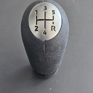 May include: A black gear shift knob with a silver top. The top has a standard 5-speed gear pattern with the numbers 1, 2, 3, 4, 5 and the letter R.