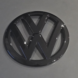 Könnte beinhalten: Ein glänzend schwarzes Volkswagen-Emblem mit dem ikonischen "VW"-Logo. Das Emblem ist kreisförmig mit einem erhabenen, dreidimensionalen Design. Die schwarze Oberfläche sorgt für eine elegante, moderne Ästhetik.