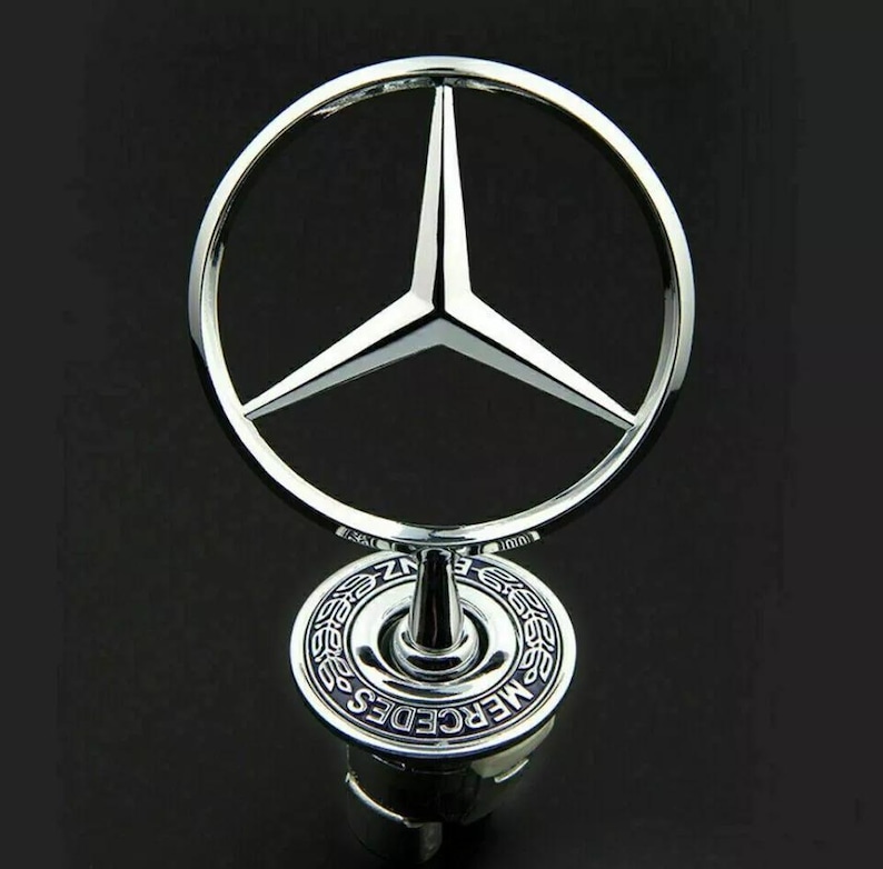 May include: A chrome-finished Mercedes-Benz hood ornament. The emblem features a three-pointed star within a circle, mounted on a base with the brand name. The ornament is highly reflective against a dark background.