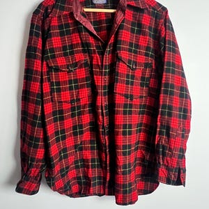 Peut inclure: Une chemise en flanelle à carreaux rouges, noirs et jaunes avec deux poches de poitrine. La chemise a une fermeture à boutons et un col bordeaux. La chemise est suspendue à un cintre en bois.