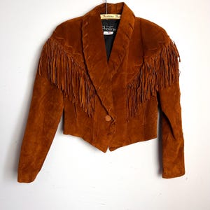 May include: A brown suede jacket with fringe detailing on the chest and sleeves. The jacket has a notched collar, a single button closure, and a tailored fit. The label reads "Fashion Junx".