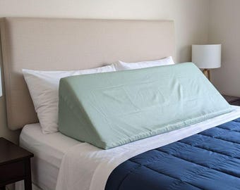 May include: A sage green wedge pillow with a smooth fabric cover positioned on a bed. The pillow has a distinct triangular shape and is placed horizontally across the mattress in front of two white pillows against a beige headboard.