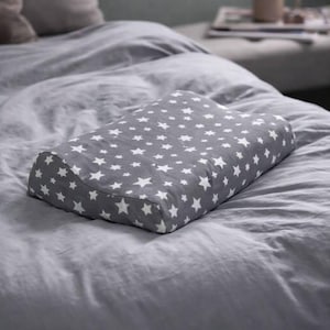 May include: A rectangular contoured pillow featuring a grey fabric cover with a repeating pattern of small white stars. The pillow has a distinct curved shape designed for neck support and is resting on a grey bed sheet with a soft texture.