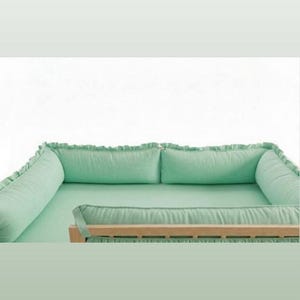 Puede incluir: Un nido para bebé verde menta con estructura de madera. El nido presenta un interior suave y acolchado y un borde con volantes. El diseño incluye una base rectangular y un refuerzo circundante para mayor comodidad y seguridad.