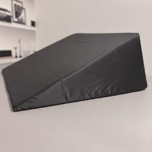 Puede incluir: Una almohada de cuña de espuma negra con una funda de tela lisa y mate. La almohada presenta una forma triangular inclinada con una base plana y una superficie superior en pendiente, diseñada para proporcionar elevación y apoyo.