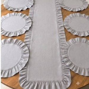 May include: A light gray table runner and six matching circular placemats. Each piece features a solid light gray center and a gathered, ruffled fabric border. The set is arranged symmetrically, with the runner centered between the placemats.