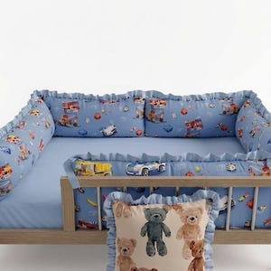 Könnte beinhalten: Ein hellblaues Kinderbett-Set mit einem Muster aus bunten Spielzeugautos, Lastwagen und Bausteinen, komplett mit Rüschenkanten. Ein quadratisches Dekokissen mit cremefarbenem Hintergrund und einem Aufdruck verschiedener brauner und grauer Teddybären.