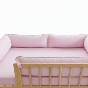 Könnte beinhalten: Ein hellrosa Kinderbett-Set bestehend aus einem Spannbettlaken und vier gepolsterten Nestchen, die den Innenrand eines hellen Holzgitterbetts mit vertikalen Stäben auskleiden und ein weiches, einheitliches pastellfarbenes Erscheinungsbild bieten.