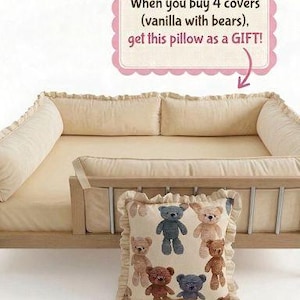 May include: A beige pillow with a ruffle trim featuring a pattern of teddy bears in various colors. The pillow is displayed in front of a light-colored bed frame with matching bedding. The image includes text.