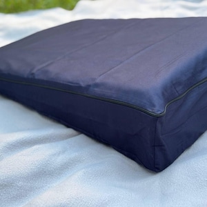 May include: A navy blue wedge-shaped cushion featuring a smooth fabric cover and a thin black piped edge detail along the perimeter. The cushion has a rectangular base that tapers toward the top, creating a sloped surface for support.