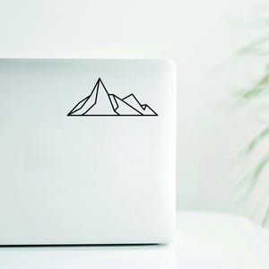Puede incluir: Una pegatina negra con una cordillera geométrica en un portátil blanco. La pegatina presenta un dibujo lineal simple de varios picos montañosos. El portátil está sobre una superficie blanca, con una planta verde borrosa en el fondo.