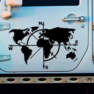 Op de afbeelding: Een zwart-witte wereldkaart sticker met een kompasroos op een witte autodeur. De kompaspunten zijn gemarkeerd met N, S, E en W.