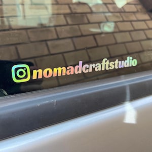 Könnte beinhalten: Ein holografischer Aufkleber mit dem Instagram-Logo und dem Text "nomadcraftstudio". Der Aufkleber hat einen Farbverlauf in Regenbogenfarben. Er ist an einer Autoscheibe angebracht.