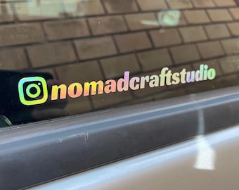 May include: A holographic sticker with the Instagram logo and the text "nomadcraftstudio". The sticker has a gradient of colors, including yellow, green, pink, and purple. It is affixed to a car window.