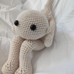 Peut inclure: Poupée au crochet beige clair. Elle a une tête ronde avec de grands yeux noirs, de petites oreilles et de longs membres fins. La poupée est assise, les jambes pliées et les pieds posés. Fabriquée en fil doux, elle est faite à la main.