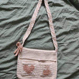 May include: A cream-colored crochet shoulder bag with a long strap. The bag features two front pockets with brown heart and bunny designs. A small brown crochet figure hangs from the side. The bag is set against a green fabric background.