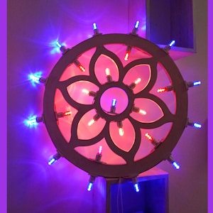 Può includere: Un oggetto decorativo a forma di ruota illuminata con un design floreale. La ruota è illuminata con luci rosse e bianche al centro e luci blu attorno al perimetro. Lo sfondo è un gradiente di viola e rosa.