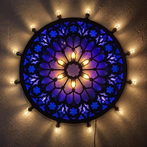 May include: A circular wall light fixture featuring a black frame with a blue stained glass-style insert. The design includes intricate floral and geometric cutouts. Small warm-toned light bulbs are mounted around the outer edge and center.