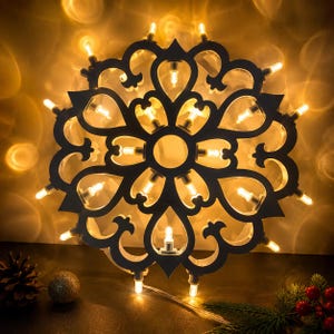 Può includere: Una cornice decorativa circolare di colore scuro caratterizzata da un intricato motivo ritagliato di forme simmetriche simili a fiori. La cornice è illuminata da una serie di piccole luci LED dai toni caldi posizionate nei ritagli.