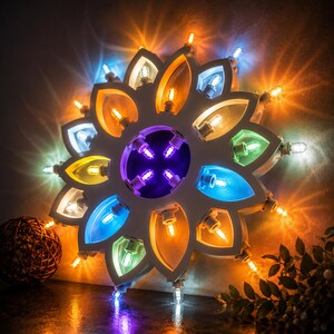 Può includere: Una lampada bianca a forma di fiore con lampadine colorate. I petali sono illuminati con luci arancioni, blu, verdi e gialle. Il centro ha luci viola. La lampada è progettata per essere una luce decorativa.
