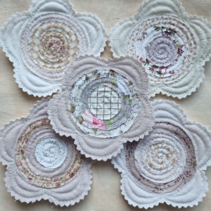 Puede incluir: Cinco posavasos de tela blanca con un dise&ntilde;o circular en tonos beige, marr&oacute;n y gris. Los posavasos est&aacute;n hechos de tela en capas y tienen un borde festoneado.