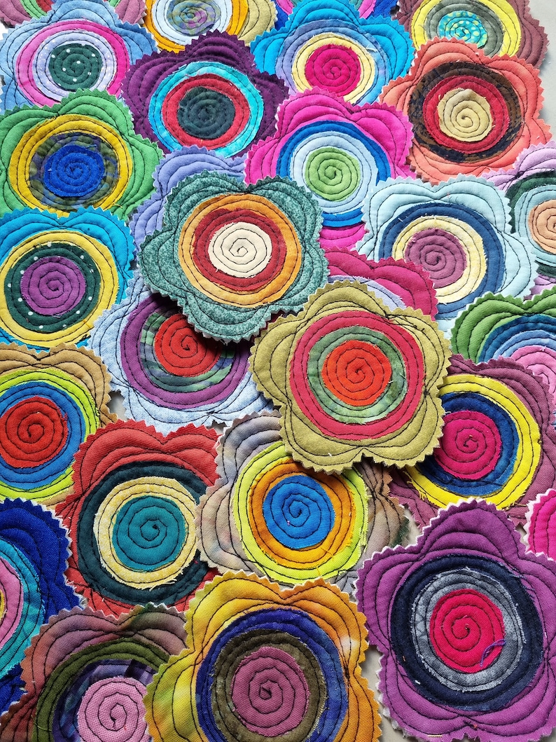 K&ouml;nnte beinhalten: Eine Sammlung bunter Stoffuntersetzer mit Blumenform. Jeder Untersetzer zeigt ein Spiralmuster in verschiedenen Schattierungen von Rot, Blau, Gr&uuml;n, Gelb und Lila.