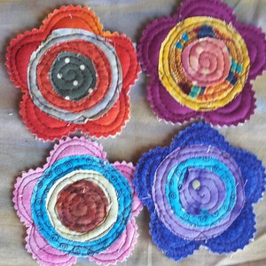 K&ouml;nnte beinhalten: Vier handgefertigte Stoff-Untersetzer in den Farben Rot, Lila, Rosa und Blau. Jeder Untersetzer hat ein kreisf&ouml;rmiges Design mit konzentrischen Kreisen und Spiralen.