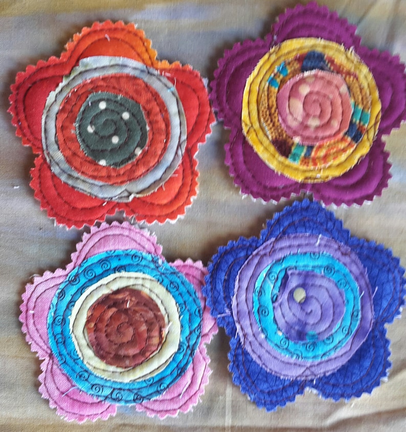 K&ouml;nnte beinhalten: Vier handgefertigte Stoff-Untersetzer in den Farben Rot, Lila, Rosa und Blau. Jeder Untersetzer hat ein kreisf&ouml;rmiges Design mit konzentrischen Kreisen und Spiralen.