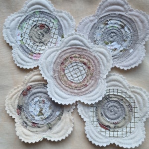 Puede incluir: Cinco posavasos de tela hechos a mano con forma de flores. Cada posavasos presenta un patr&oacute;n diferente de c&iacute;rculos conc&eacute;ntricos en tonos grises, blancos y rosas.