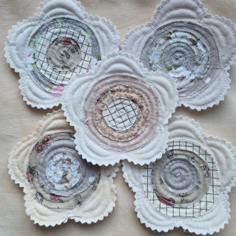 Puede incluir: Cinco posavasos de tela hechos a mano con forma de flores. Cada posavasos presenta un patr&oacute;n diferente de c&iacute;rculos conc&eacute;ntricos en tonos grises, blancos y rosas.