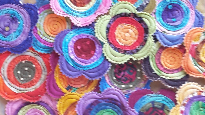 K&ouml;nnte beinhalten: Eine Sammlung bunter Stoffblumen mit mehreren Stofflagen in Blaut&ouml;nen, Rot, Orange, Gr&uuml;n und Lila. Die Blumen sind zusammengen&auml;ht und haben einen einzigartigen, handgefertigten Look.