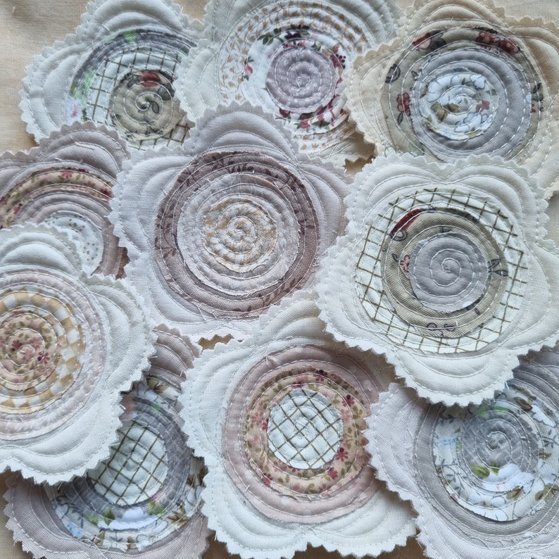 Puede incluir: Un juego de 10 posavasos de tela hechos a mano con una variedad de patrones en tonos blancos, beige y marrones. Los posavasos son circulares y tienen una textura acolchada.